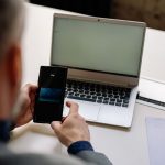 Profissional usa o celular para realizar um pagamento digital ao lado de um laptop em um escritório.